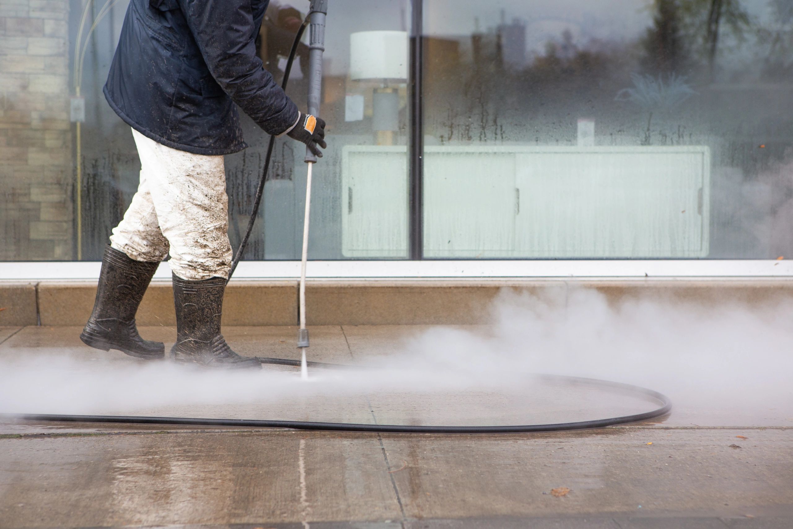 Worker power washing the sidewalk wearing black rubber.